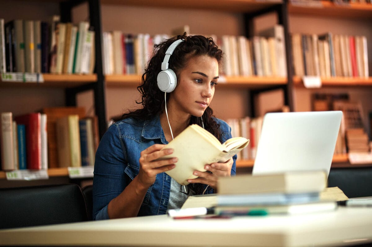 Female student with headphones holding an open book and looking at a laptop in a library.