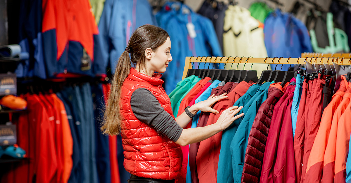 Young woman choosing winter clothes picking up down jackets on the hanger in the sports shop showing brand positioning