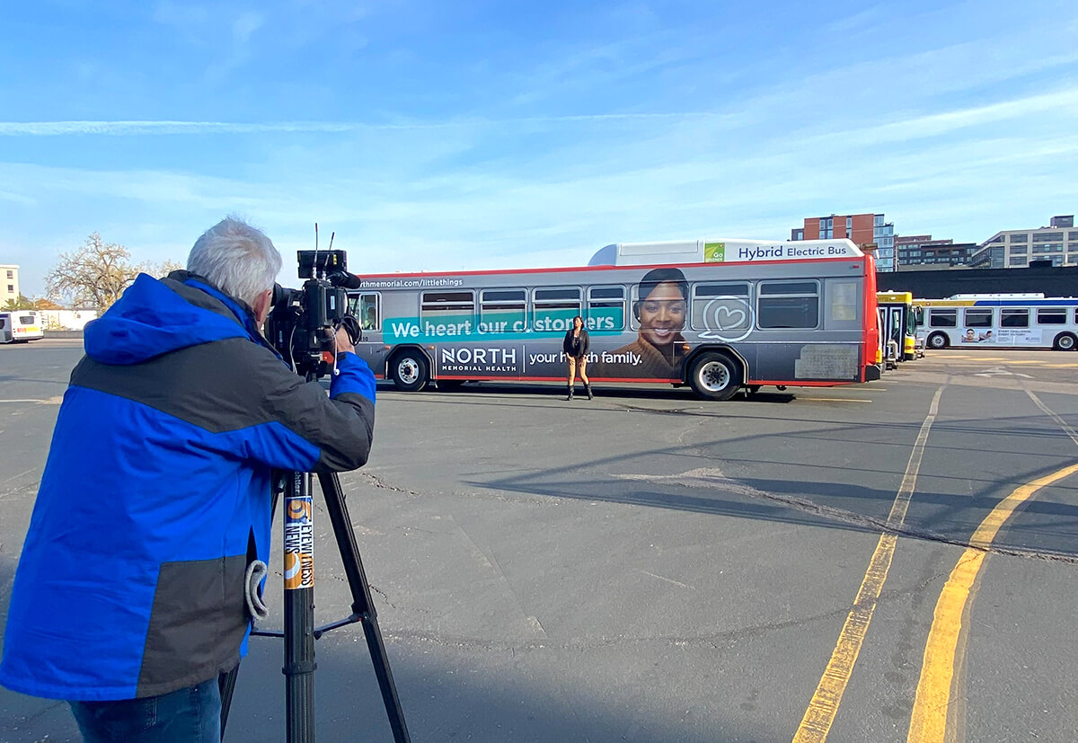 Behind the scenes photo of a videographer and featured woman in front of a bus wrapped in branded graphics.