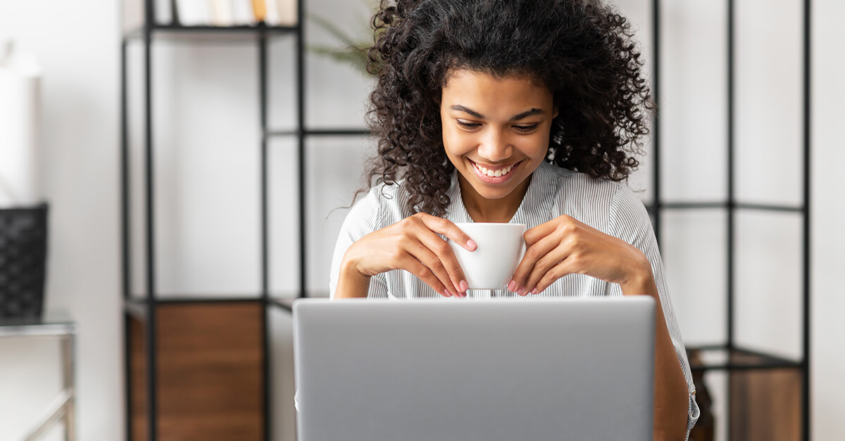 Smiling young cheerful African American woman with Afro hairstyle enjoying morning coffee while watching video news on the laptop, sitting at the desk in the home office, businesswoman having a break.